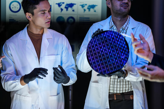 Two researchers inspect the die layout on a wafer while a gloved hand steadies the edge of the disc inside a dimly lit research and development laboratory late in the working day