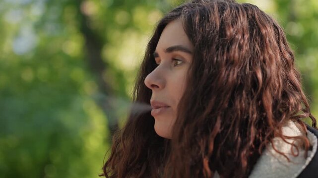 Close up view shows young adult female in outdoor setting. Woman holds lit cigarette between fingers with long wavy brown hair and casual attire visible. Background is softly blurred