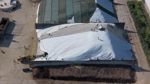 Aerial view of agricultural machinery and workers handling stored forage inside large silage pits, showing mechanized farm operations and livestock feed preparation in Italy.