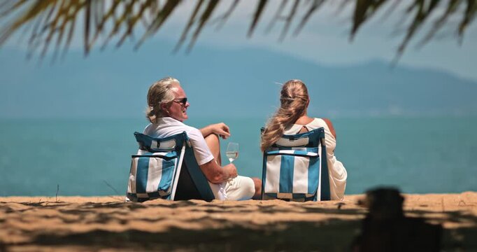 Thailand travel destination. Mature couple enjoying a luxury resort vacation, sitting in beach chairs under palm shade, drinking white wine, and watching clear turquoise ocean. Samui island paradise