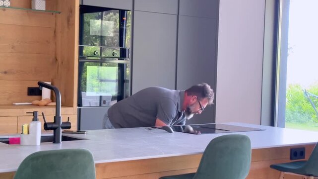 Middle-aged man with a beard and glasses preparing a meal in a contemporary kitchen, stirring food in a pot on an electric induction hob while waiting for it to cook by the window