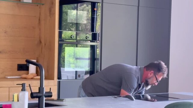 Bearded man with glasses wearing a polo shirt preparing a meal in a contemporary kitchen, placing a pot on an induction hob, stirring the contents, and waiting for it to cook