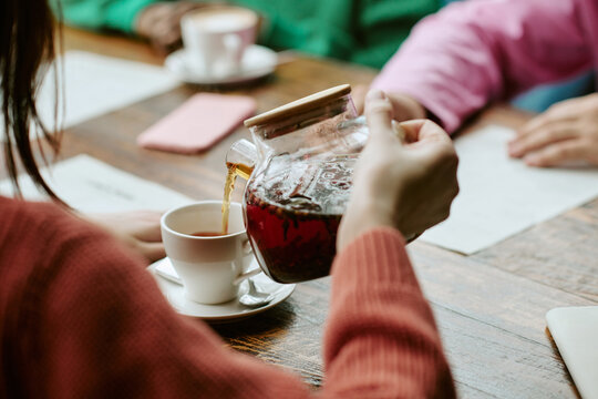 Cropped shot of young adult woman pouring hot herbal tea into cup while meeting multiethnic friends in cafe. Useful for beverage branding, social lifestyle campaigns and leisure advertising