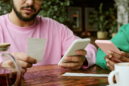 Multiethnic young adult friends sitting in cafe, checking paper bill receipt and using smartphones for splitting payment. Suitable for mobile banking, finance apps, fintech marketing