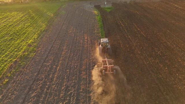 Aerial view of tractor with harrow cultivating soil and raising dust next to green field at sunset