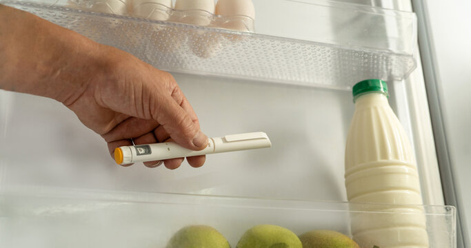 A man's hand places an injection pen containing medication into home refrigerator next to food items. Concept of home-based treatment, medication storage, and use of drugs requiring refrigeratio