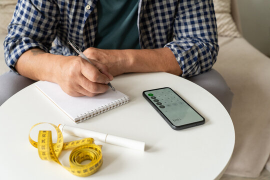 Man at home records weight-related data in notebook, pen injector, measuring tape, smartphone with calculator lie nearby. Concept of metabolic health, calorie counting, self-administered medication