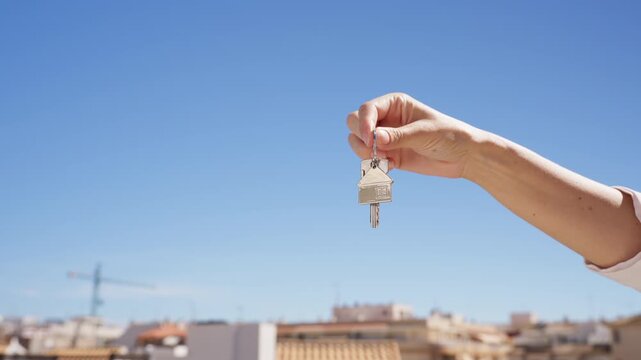 Close-up of female hand hold new home keys with house-shaped keychain against clear blue sky, symbolizing property purchase, home ownership, moving into new apartment and real estate investment