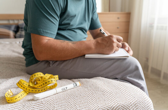 Mature overweight man sits on bed, writes in notepad. Tracks your health and weight loss progress. Nearby are measuring tape and an injection pen with inscription GLP