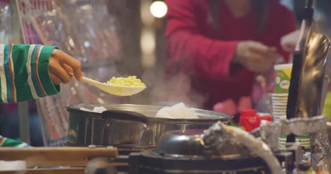 A street food vendor scoops steamed sweet corn kernels with a spoon from a pot at a Thai night market stall.