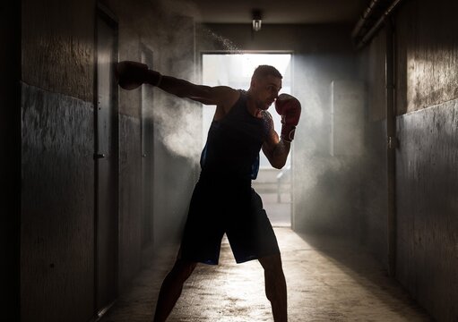 Dramatic silhouette of a strong male boxer throwing a punch with flying dust in a dark underground gym corridor, or intense fighter shadowboxing for competitive martial arts fitness lifestyle.