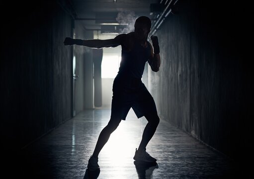 Dramatic silhouette of a strong male boxer throwing a punch with flying dust in a dark underground gym corridor, or intense fighter shadowboxing for competitive martial arts fitness lifestyle.