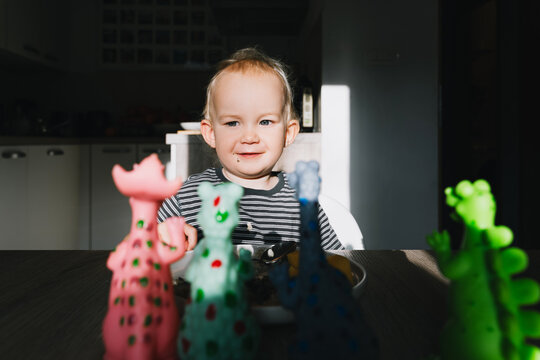 Cute toddler boy eats porridge with mango while playing with colorful toy dinosaurs at a home dining table. Authentic childhood lifestyle concept showing playful everyday mealtime moments.