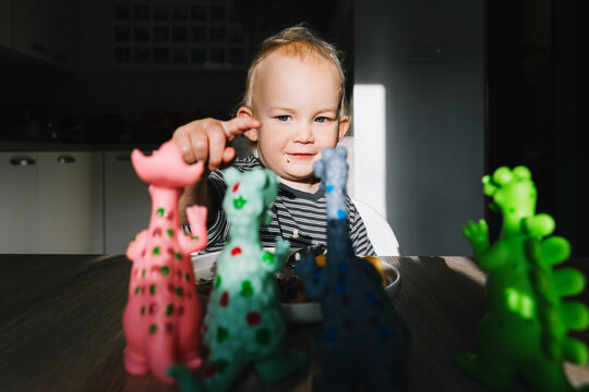 Cute toddler boy eats porridge with mango while playing with colorful toy dinosaurs at a home dining table. Authentic childhood lifestyle concept showing playful everyday mealtime moments.