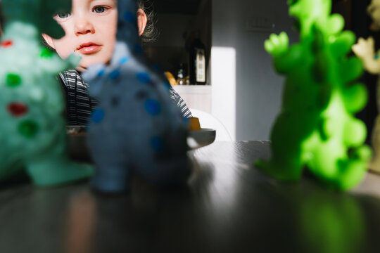 Cute toddler boy eats porridge with mango while playing with colorful toy dinosaurs at a home dining table. Authentic childhood lifestyle concept showing playful everyday mealtime moments.