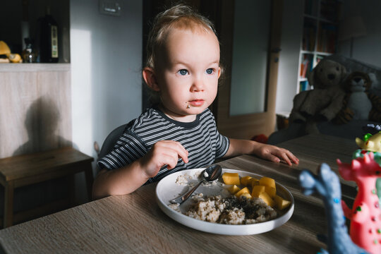 Cute toddler boy eats porridge with mango while playing with colorful toy dinosaurs at a home dining table. Authentic childhood lifestyle concept showing playful everyday mealtime moments.