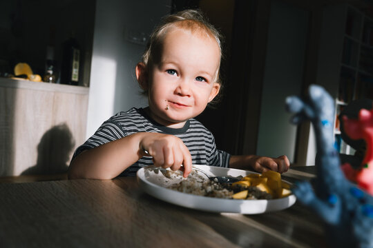 Cute toddler boy eats porridge with mango while playing with colorful toy dinosaurs at a home dining table. Authentic childhood lifestyle concept showing playful everyday mealtime moments.