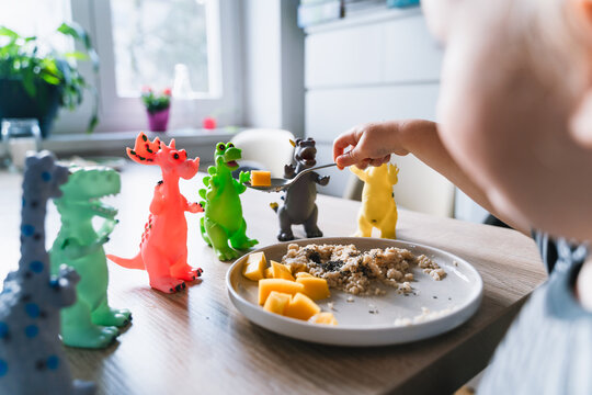 Cute toddler boy eats porridge with mango while playing with colorful toy dinosaurs at a home dining table. Authentic childhood lifestyle concept showing playful everyday mealtime moments.