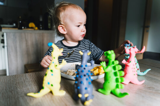 Cute toddler boy eats porridge with mango while playing with colorful toy dinosaurs at a home dining table. Authentic childhood lifestyle concept showing playful everyday mealtime moments.