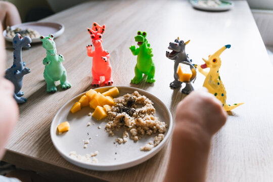 Cute toddler boy eats porridge with mango while playing with colorful toy dinosaurs at a home dining table. Authentic childhood lifestyle concept showing playful everyday mealtime moments.