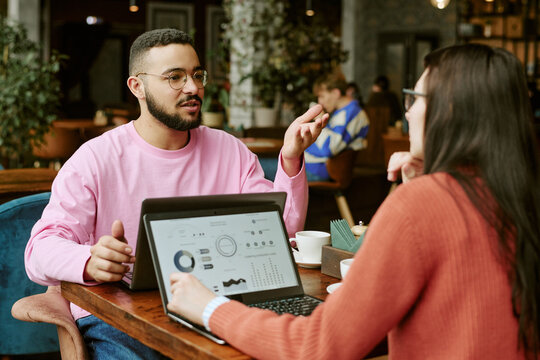 Young Biracial man and Caucasian woman discussing business data on laptops in cafe. Image suitable for remote work, startup collaboration, and digital marketing campaigns