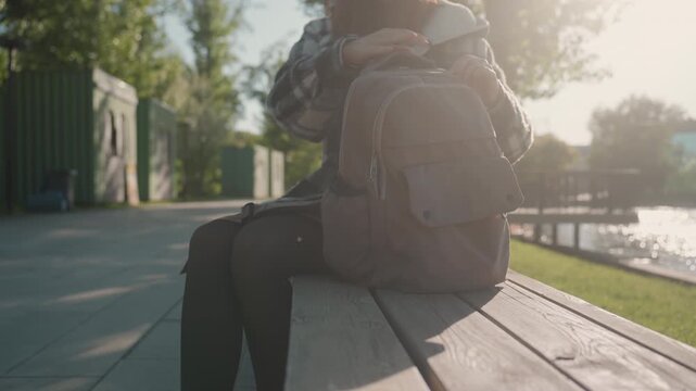 Young woman wearing plaid jacket is seated outdoors near calm waters. Woman interacts with open pink backpack on wooden bench adjusting its contents while surrounded by greenery and distant