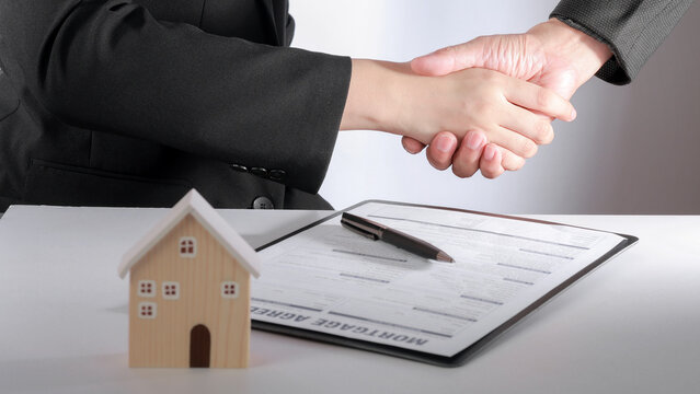 Confident business handshake marks completed housing finance process beside estate contract model house representing successful deal agreement