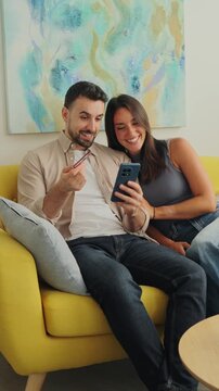 A couple sits together on a yellow sofa, smiling as they look at a phone and hold a card. The scene features a light, airy living room with a blue abstract painting and natural daylight, suggesting a