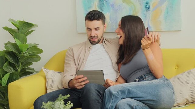 A cheerful couple sits together on a bright yellow sofa, smiling while browsing on a tablet and waving a credit card. Natural daylight and a colorful abstract wall art add warm tone and focus on