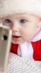 Happy baby girl in red Santa costume laughing during interactive video call. Tiny kid looking at smartphone screen with cheerful holiday child emotion. Virtual Christmas family greetings © Marina Demidiuk