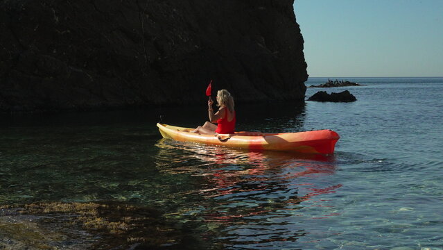 Kayaking vacation sea woman paddling kayak near dark rugged cliff silhouette on crystal clear blue ocean water.