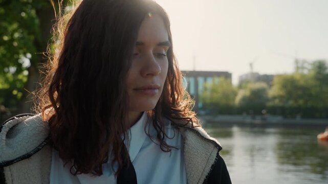 Woman is shown from side as woman looks out over calm waters on sunny day. Her long hair catches light and her clothing suggests casual outdoor attire. background includes blurred greenery