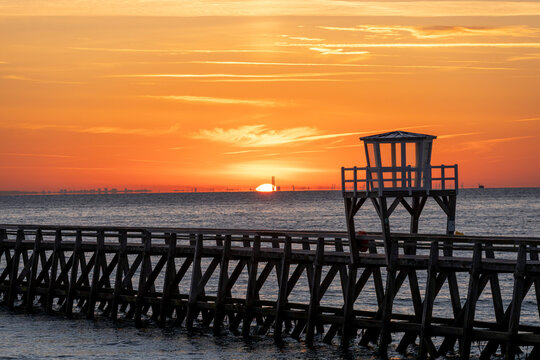 Luc-Sur-Mer, France - 08 08 2026:  Panoramic view of a wooden pier in front of the beach, sea, a colorful sunrise and Le Havre city far away