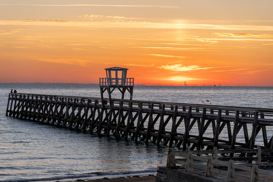 Luc-Sur-Mer, France - 08 08 2026:  Panoramic view of a wooden pier in front of the beach, sea, a colorful sunrise and Le Havre city far away
