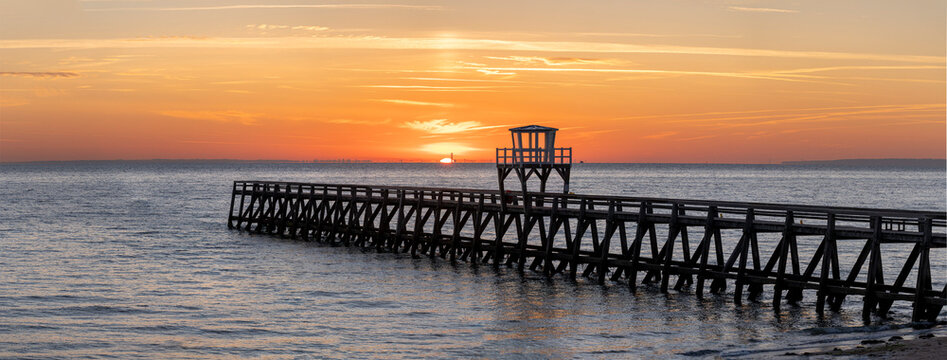 Luc-Sur-Mer, France - 08 08 2026:  Panoramic view of a wooden pier in front of the beach, sea, a colorful sunrise and Le Havre city far away
