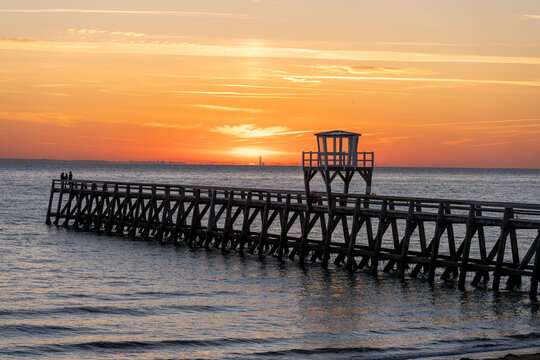Luc-Sur-Mer, France - 08 08 2026:  Panoramic view of a wooden pier in front of the beach, sea, a colorful sunrise and Le Havre city far away