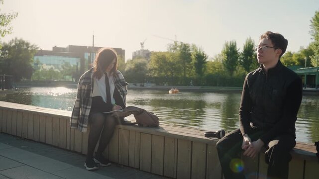 Young woman sits on wooden bench near lake or pond writing notes while looking at paper. man is seated next to her facing forward with hands clasped in his lap. scene takes place outdoors