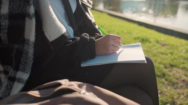 Person writes notes on paper outdoors near calm waters. Close up shows hands holding green pen over open notebook with grass and water visible in soft sunlight