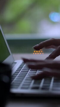 Close-up of female hands with manicured nails typing quickly on a silver laptop keyboard near a window with a blurred green background, showcasing modern remote work and digital communication