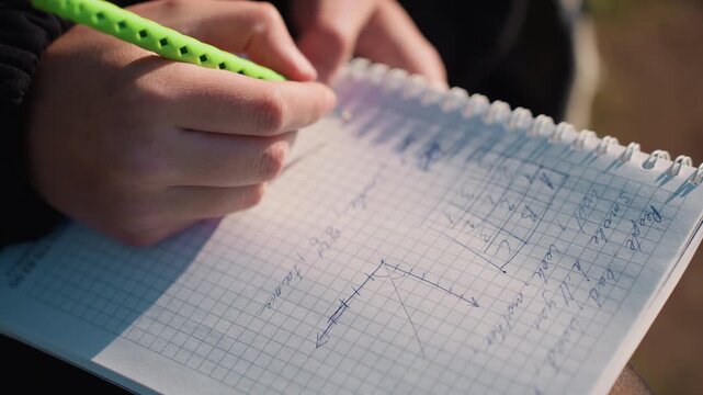Hands hold green pen over graph paper with handwritten notes and diagrams. notebook contains mathematical equations graphs and technical text written in blue ink on grid lined pages. Soft
