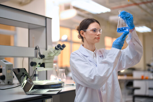 Female scientist wearing protective eyewear and gloves in a laboratory, examining a blue liquid in an erlenmeyer flask, conducting research and chemical experiments