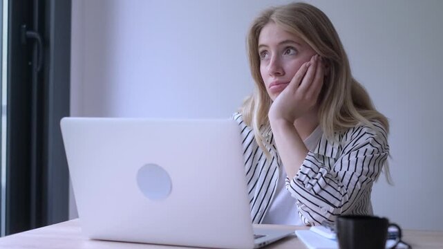 Exhausted and unmotivated young businesswoman feeling tired and bored while working on her laptop in a modern office, procrastinating and showing signs of burnout and lack of sleep