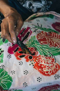 Close up of hand applying hot wax with a canting tool onto colorful patterned fabric during traditional batik making in Central Java Indonesia