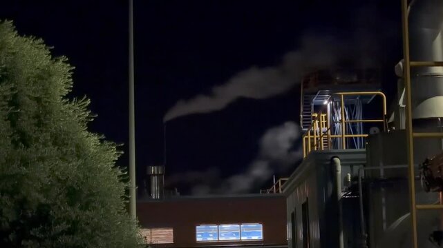 Medium shot of a chemical plant chimney emitting water vapor steam at night with an olive tree on the left and factory infrastructure on the right