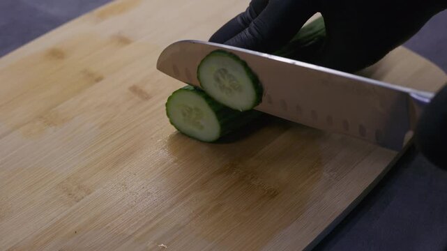 Close-up of gloved hands slicing a fresh cucumber with a kitchen knife on a wooden cutting board during food preparation.