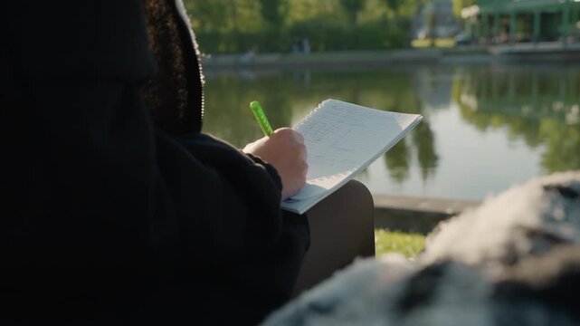 Hand writing in notebook by lake with green pen. Hand holds bright green marker while actively filling out paper set against calm outdoor water reflections
