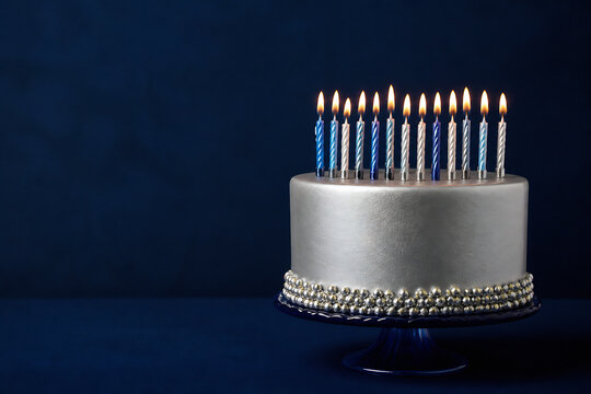 A silver birthday cake with lit candles sits on a stand against a dark blue background.