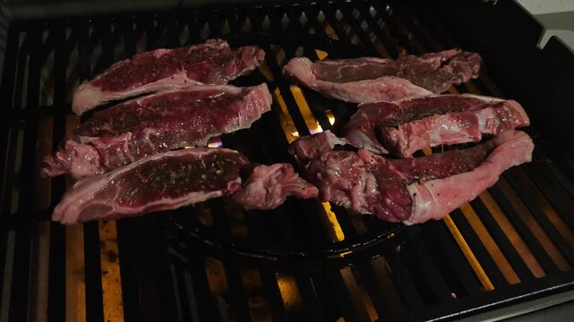 Close-up slow motion of raw red sirloin steaks grilling on a black barbecue grill in complete darkness, illuminated only by glowing flames, cinematic firelight, 4K food footage
