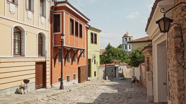 Wide view of a cobblestone street lined with National Revival architecture and the bell tower of Constantine and Helena Church in Plovdiv Old Town.