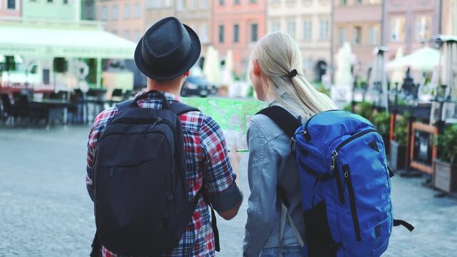 Two young travelers with backpacks checking a map together in a busy urban city square during daytime, planning their route amid outdoor cafes and buildings.